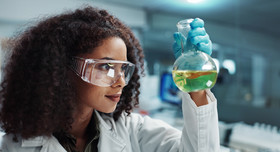 Frau mit Schutzbrille, die sich ein Reagenzglas mit grüner Flüssigkeit anschaut. Foto: Coetzee/peopleimages.com
